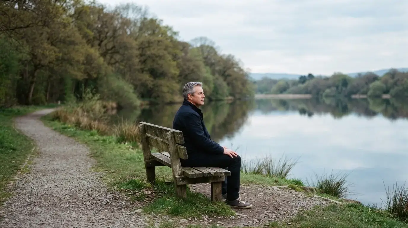 Ein Mann sitzt allein auf einer Parkbank und blickt ruhig auf einen See.